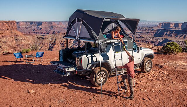A Man Helping a Woman into a Rooftop Tent on a White GMC HUMMER EV Truck Overlooking a Vast Canyon Landscape