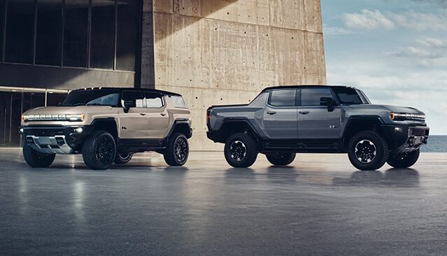 A Tan GMC Hummer EV SUV Parked Next to a Gray GMC Hummer EV Pickup Truck by a Modern Building and the Ocean