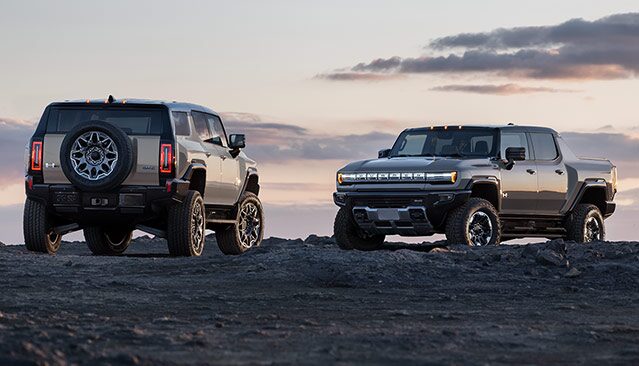 A Beige GMC HUMMER EV SUV and a Gray GMC HUMMER EV Pickup Parked on a Dirt Hill at Sunset