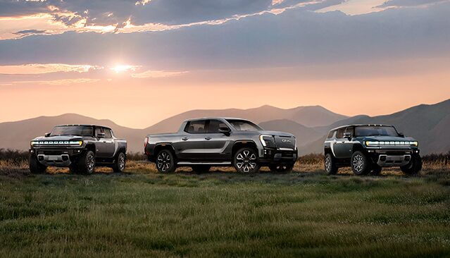 Three GMC HUMMER EV Vehicles, an SUV, a Pickup, and Another SUV, Parked in a Grassy Field with Mountains in the Background