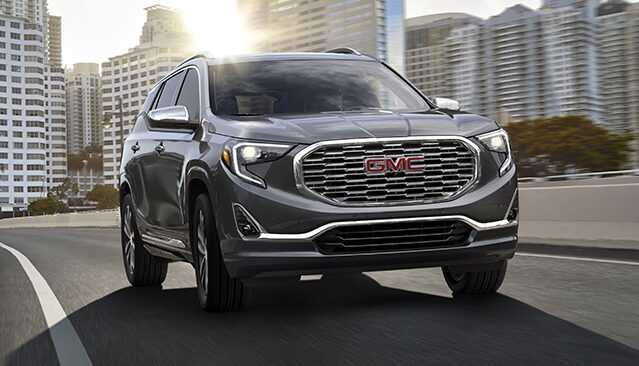 A Gray GMC Terrain SUV Driving on a Multi-Lane Highway With City Buildings in the Background