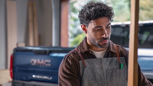 A Man in His Gargage with a Work Apron on Looking Downward and a GMC Truck Behind Him
