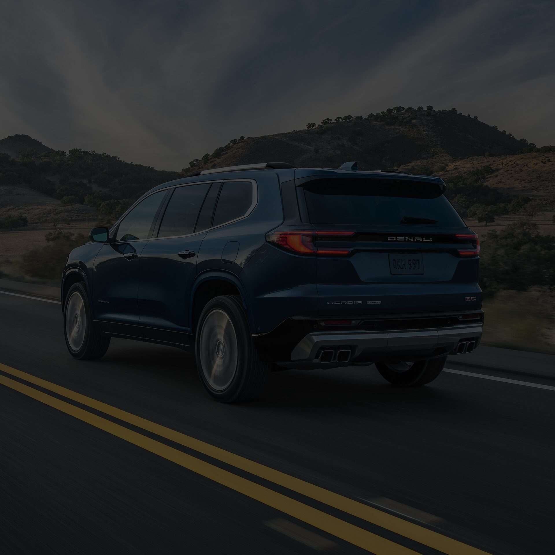 Exterior Rear View of the 2026 GMC Acadia Mid-Size SUV Driving Down a Road Lined with Mountains
