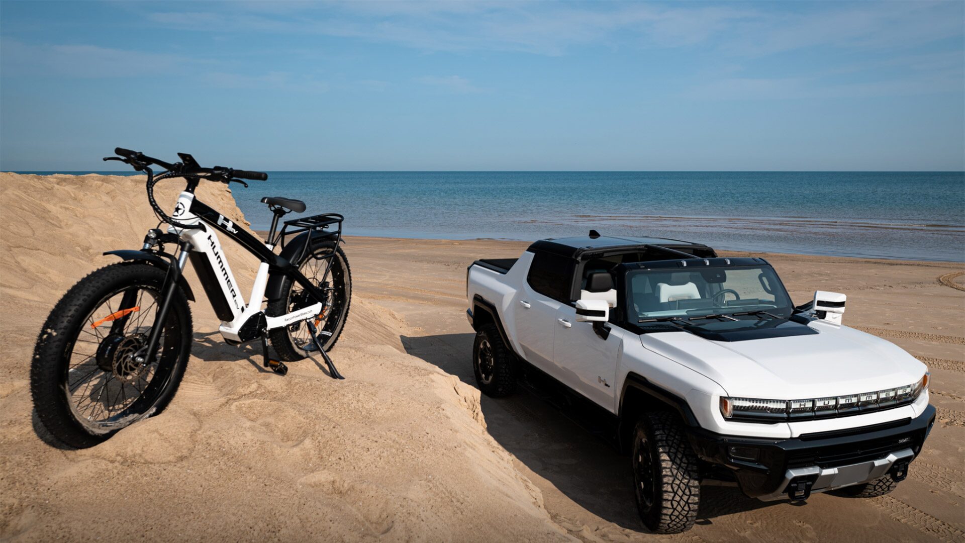 Front-Side View of the GMC HUMMER EV Pickup Parked on the Beach with the AWD Electric Bike Parked on a Hill Next to the Vehicle